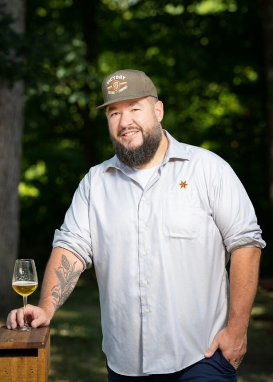 A person in a light blue shirt and gray baseball cap smiling at the camera while holding a class of beer.